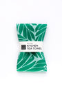 Green leaf-patterned kitchen tea towel with a label on a white background. Designed by artist Leila Simon Hayes.