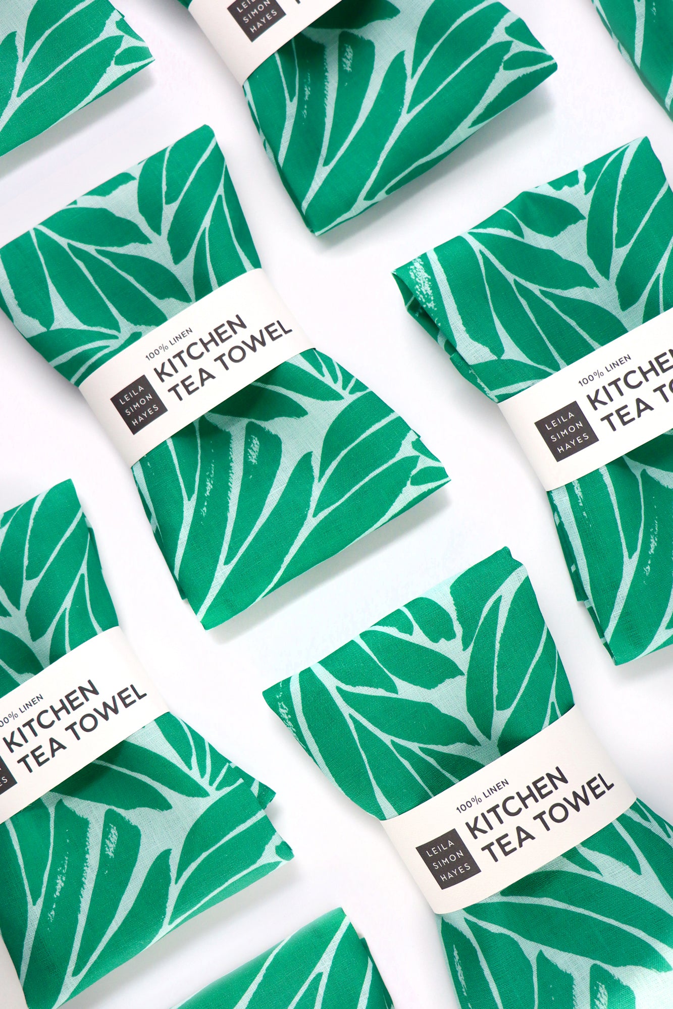 Green leaf-patterned kitchen tea towels with white labels on a white background. Designed by artist Leila Simon Hayes.