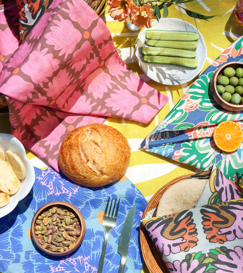 A styled picnic dining scene with colorful patterned tea towels designed by Leila Simon Hayes. The scene is sunny and includes multiple coordinated textile pieces with floral and abstract prints.