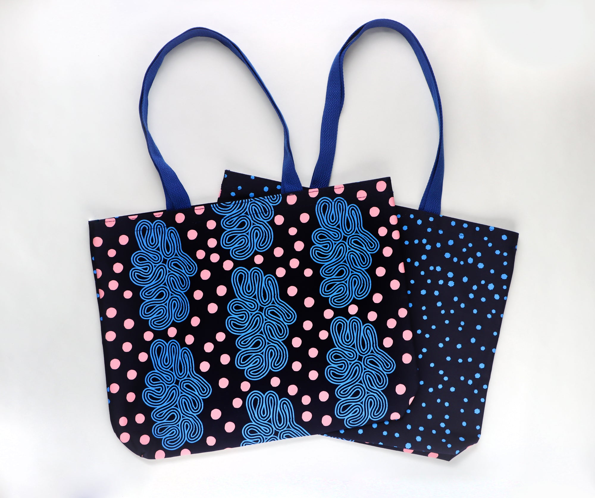 Two tote bags with blue and pink patterns on a white background. One side has blue loops design and pink dots and the other side has bright blue dots on a dark blue. Designed by artist Leila Simon Hayes.