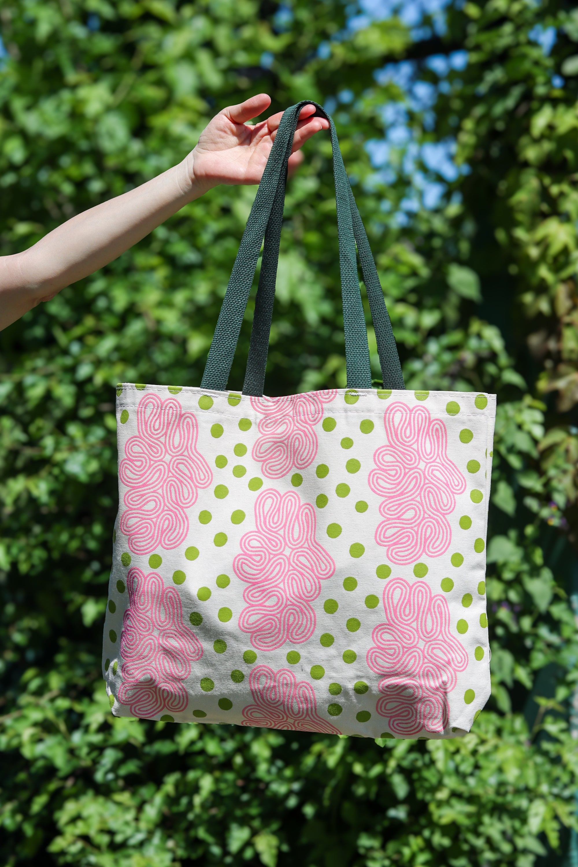 A tote bag with a pink and green abstract floral pattern on a white background, held by a person's hand against a blurred green outdoor background. Designed by artist Leila Simon Hayes.