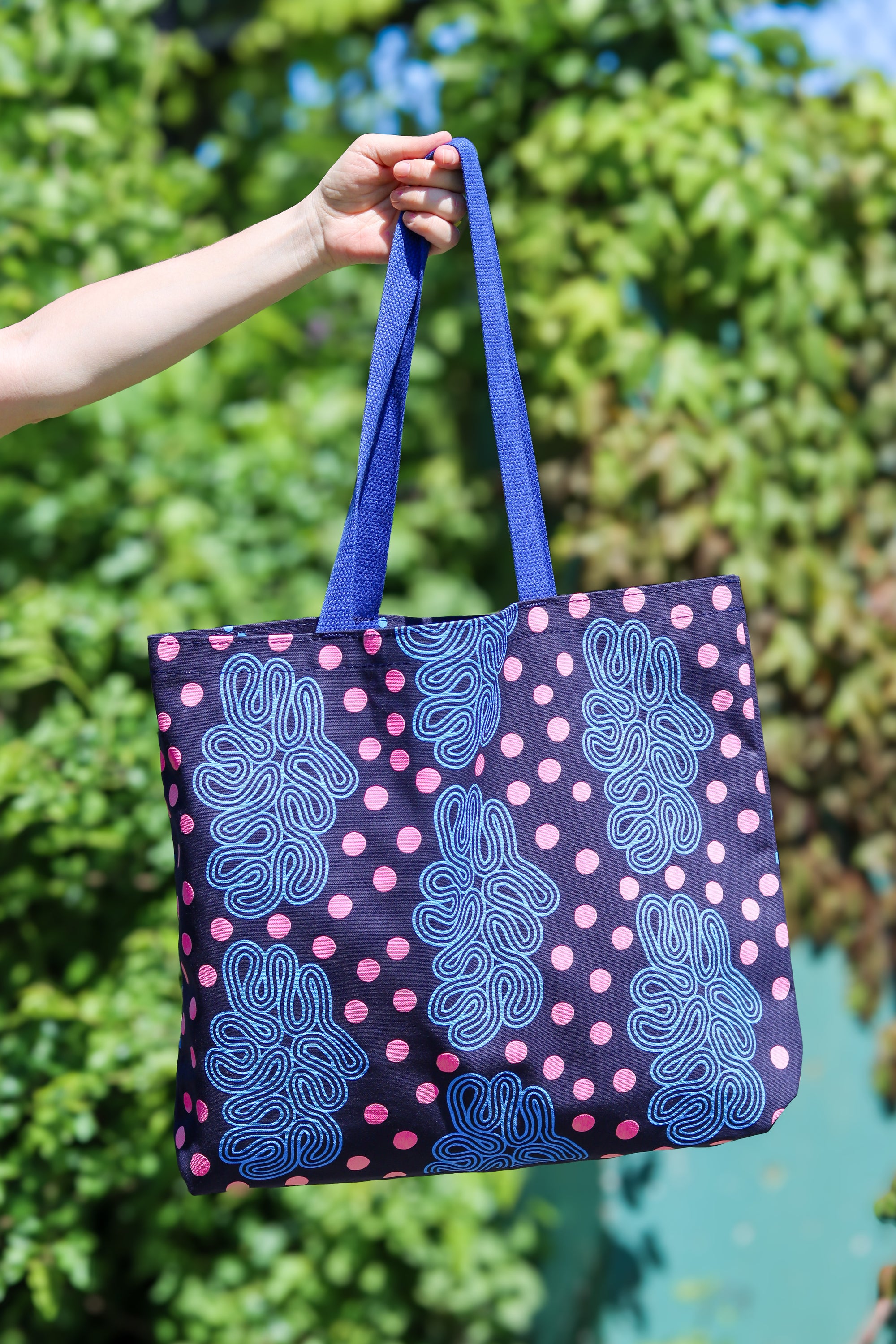 A tote bag with a navy blue base, pink polka dots, and blue abstract line patterns, held by a person's hand against a blurred green outdoor background. Designed by artist Leila Simon Hayes.