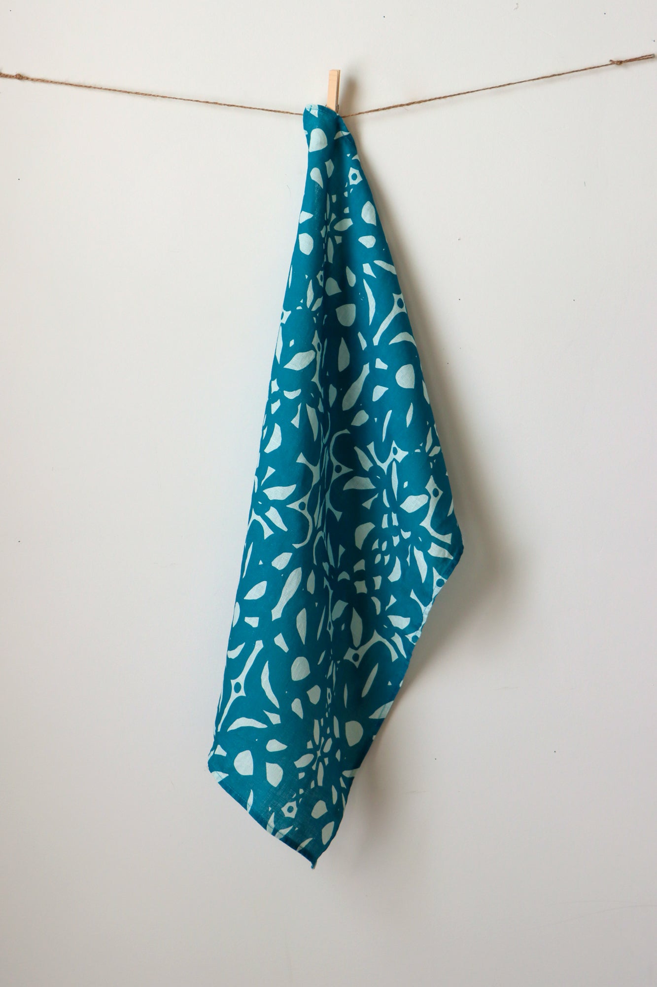 Blue tea towel with white floral pattern hanging on a line against a white background. Designed by artist Leila Simon Hayes.