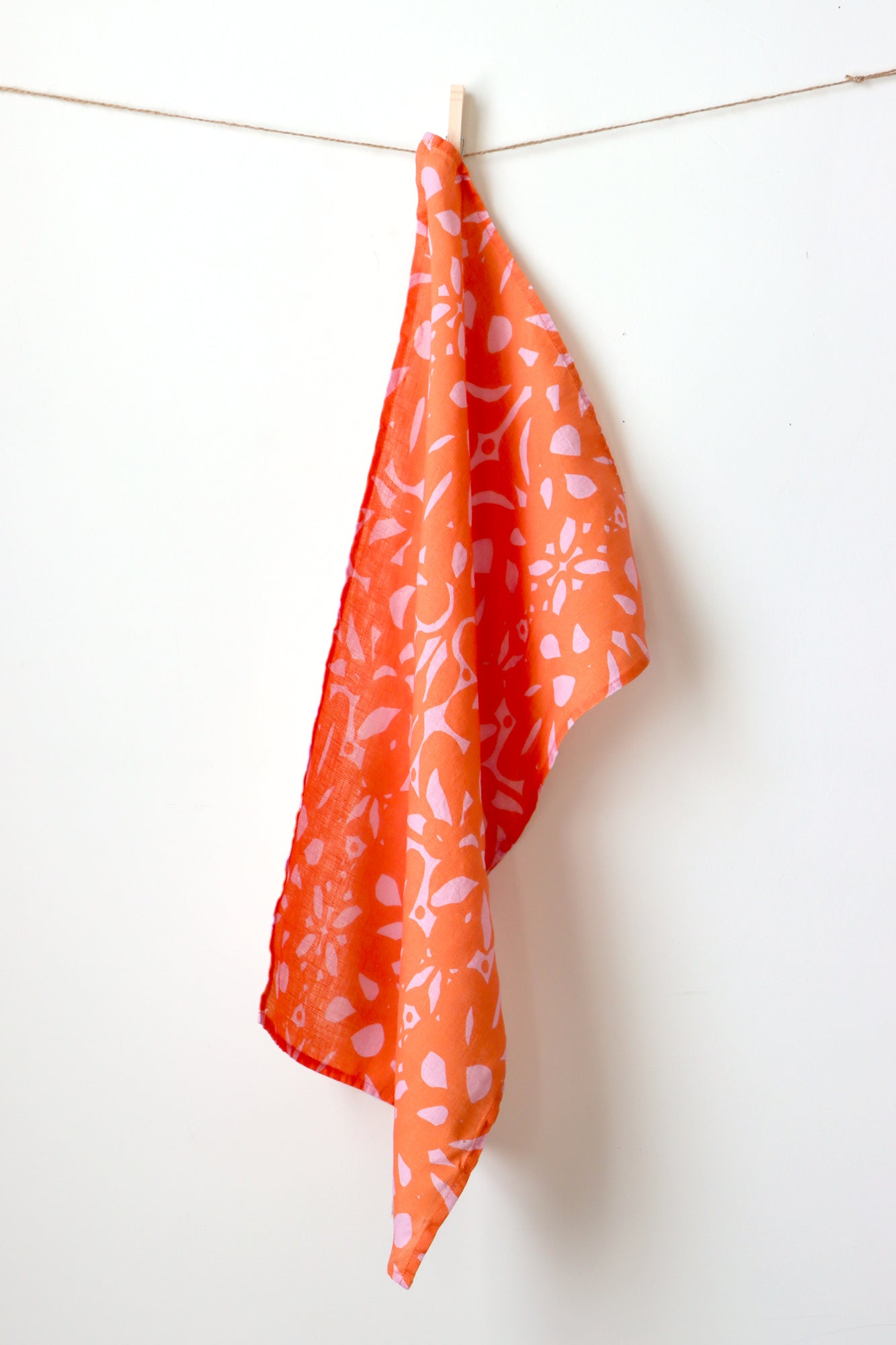 Orange towel with white floral pattern hanging on a line against a white background. Designed by artist Leila Simon Hayes.