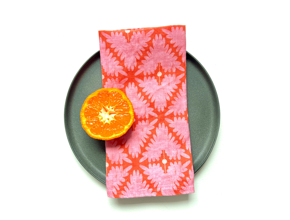 A folded pink and orange patterned cloth napkin with a geometric ikat design, displayed on a dark gray plate next to a halved orange slice on a white background. Designed by artist Leila Simon Hayes. 