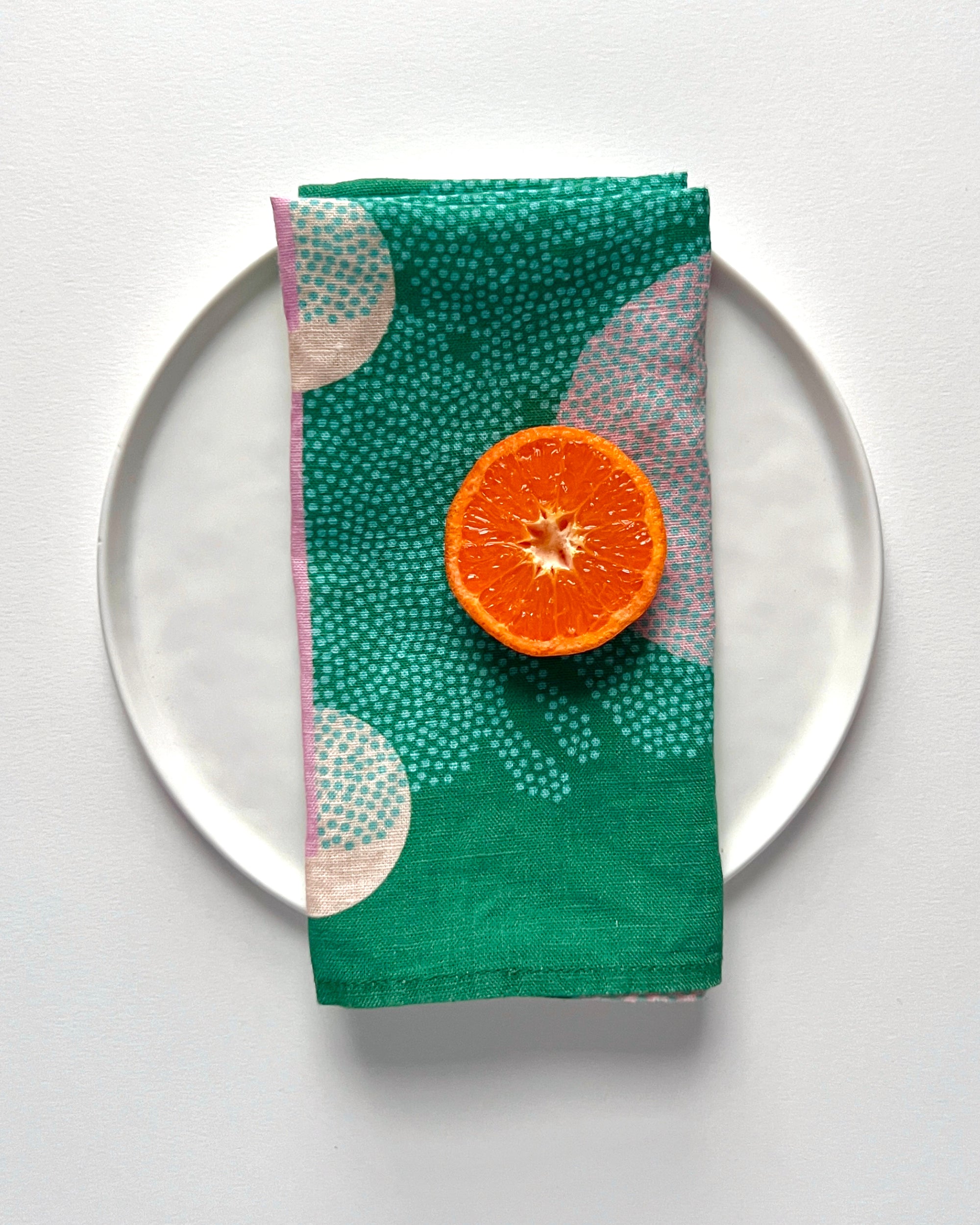 A green linen napkin with an abstract pattern is displayed on a white plate with a halved orange on top. Designed by artist Leila Simon Hayes. 