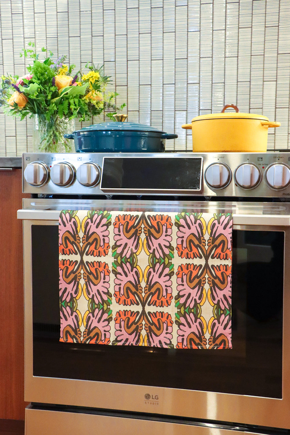 An over with a colorful patterned kitchen towel hanging on the oven door handle. A blue Dutch oven and a yellow pot sit on the stovetop, with a tiled backsplash and a vase of flowers in the background. Designed by artist Leila Simon Hayes.