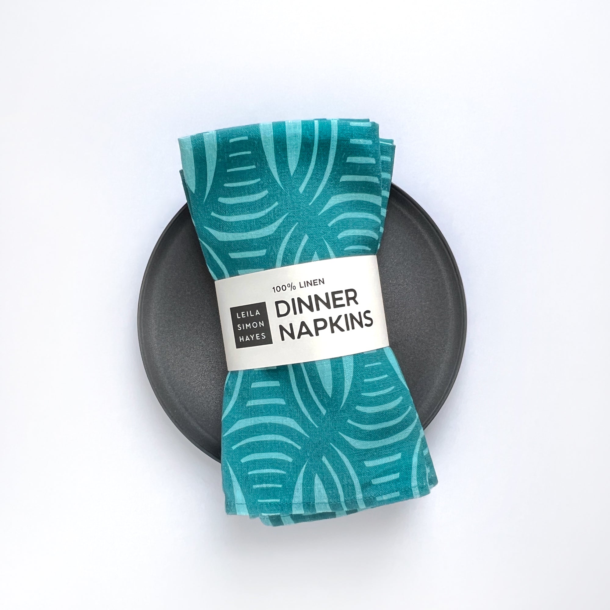 Set of teal patterned dinner napkins on a black plate with a label saying "100% linen Dinner Napkins" designed by artist Leila Simon Hayes
