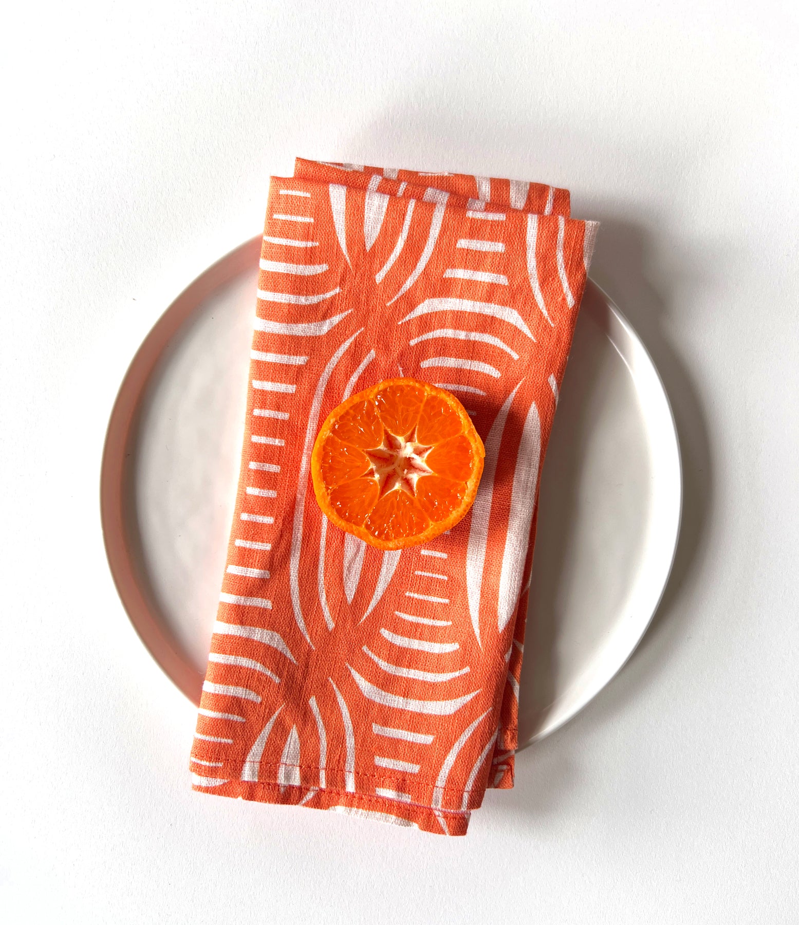 A coral colored linen napkin with a geometric pattern is displayed on a plate, with a dried orange slice on top. Designed by artist Leila Simon Hayes
