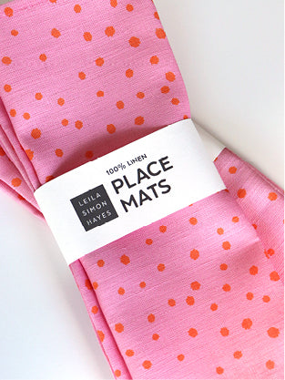 Pink place mats with organic orange dots and a visible brand label saying "100% Linen Placemats" on a white background, designed by artist Leila Simon Hayes
