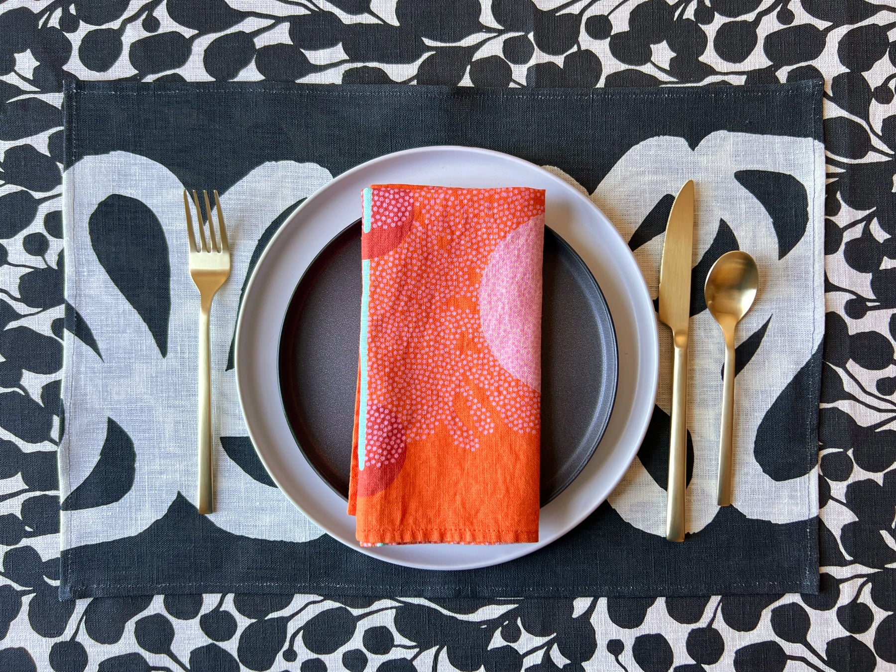 A red dinner napkin with organic dot patterns on a black and white placemat over a botanical black and white tablecloth, displayed on a table with a plate and cutlery. Designed by artist Leila Simon Hayes