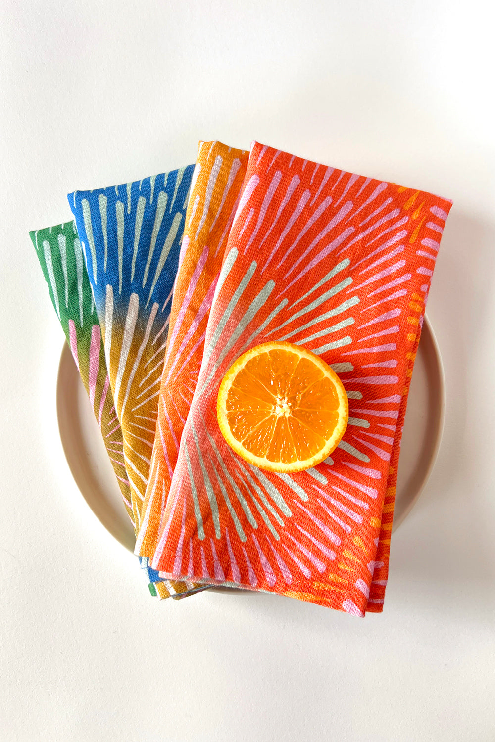 Set of four napkins with a burst design, layered on a plate and different colors, coral, orange, blue, green. Designed by artist Leila Simon Hayes. 