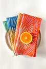 Set of four napkins with a burst design, layered on a plate and different colors, coral, orange, blue, green. Designed by artist Leila Simon Hayes. 