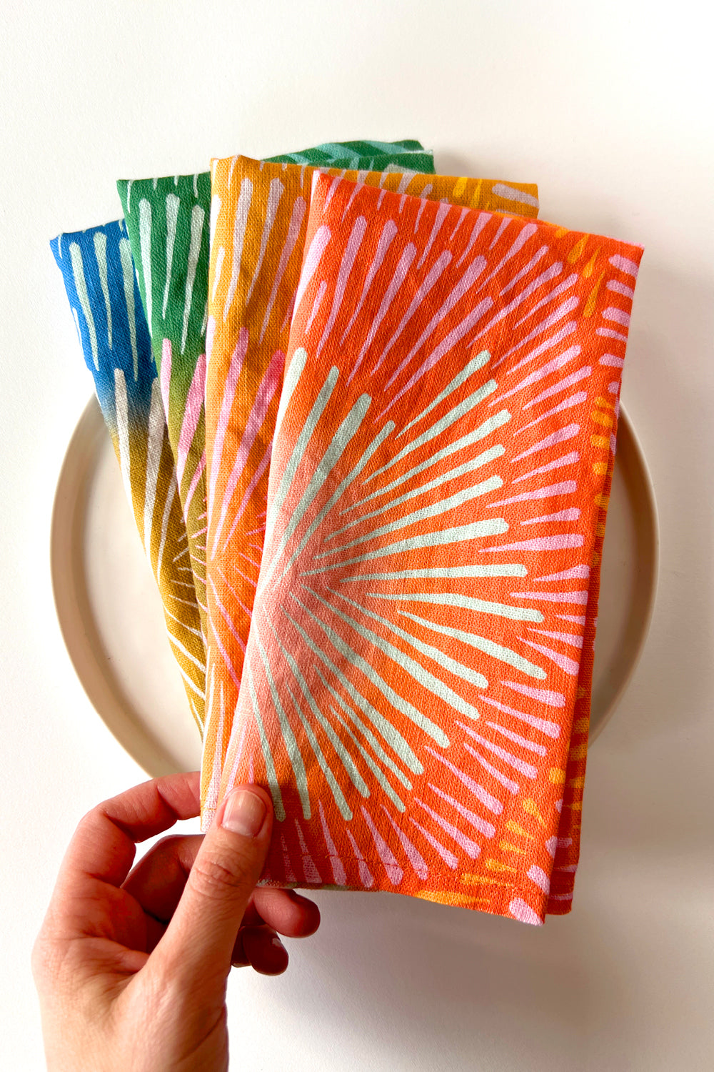 Four burst napkins in different colors layered on a white plate. Designed by artist Leila Simon Hayes. 
