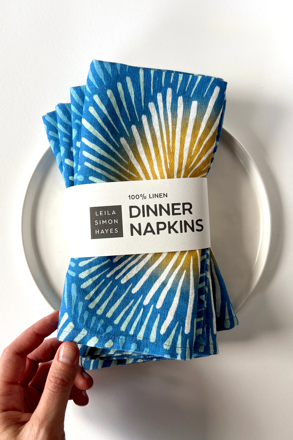 Set of four blue burst patterned dinner napkins with a label saying "100% Linen Dinner Napkins", designed by artist Leila Simon Hayes. 