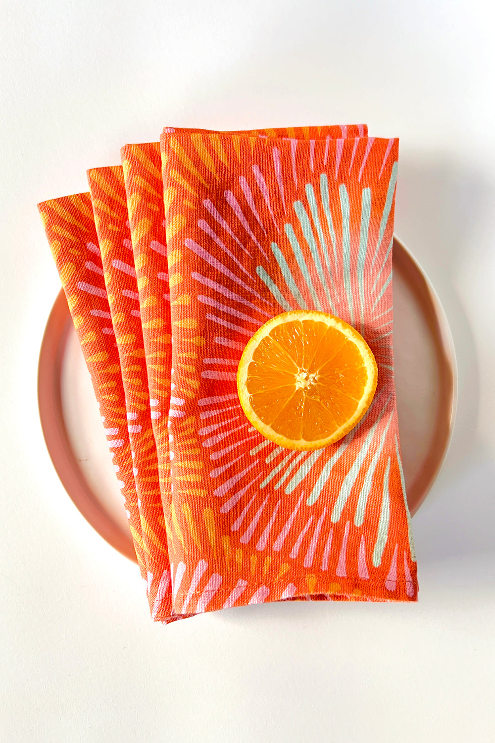 Set of four orange napkins with an abstract sunburst pattern, designed by artist Leila Simon Hayes. Napkins are layered on a white plate.