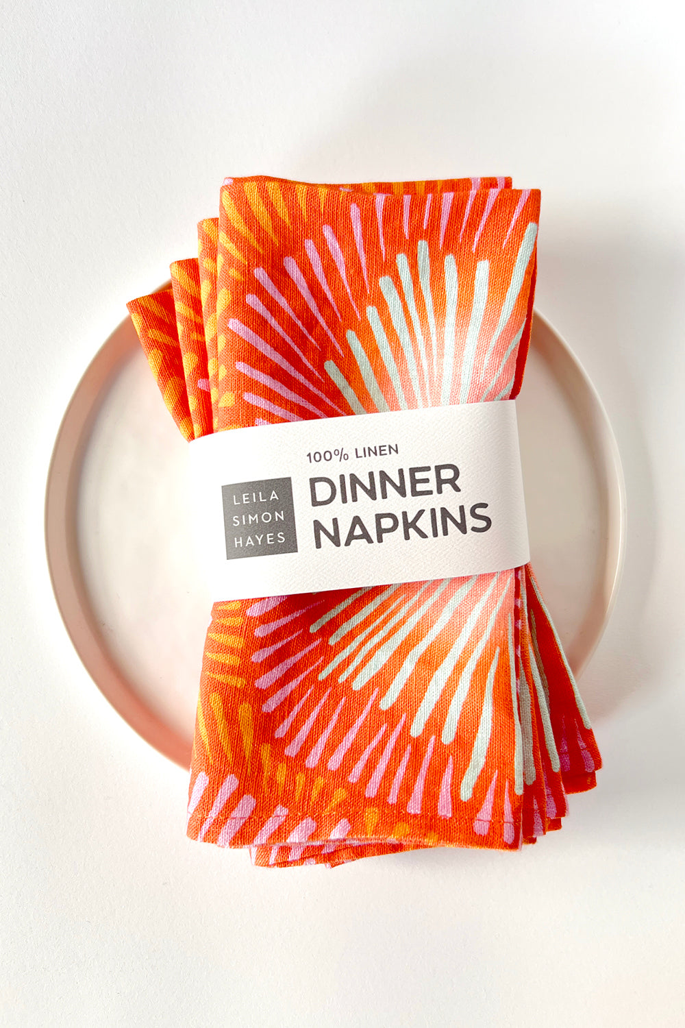 Pack of four burst patterned dinner napkins in coral with a label saying "100% Linen Dinner Napkins", designed by artist Leila Simon Hayes. 