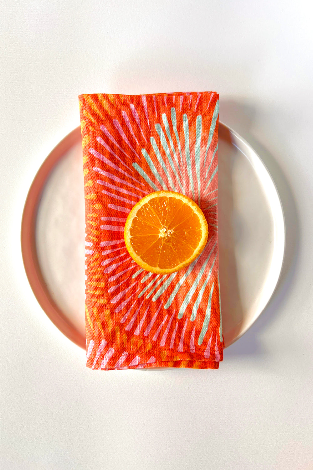 A coral colored napkin with a sunburst pattern and orange slice on a white plate. designed by artist Leila Simon Hayes. 