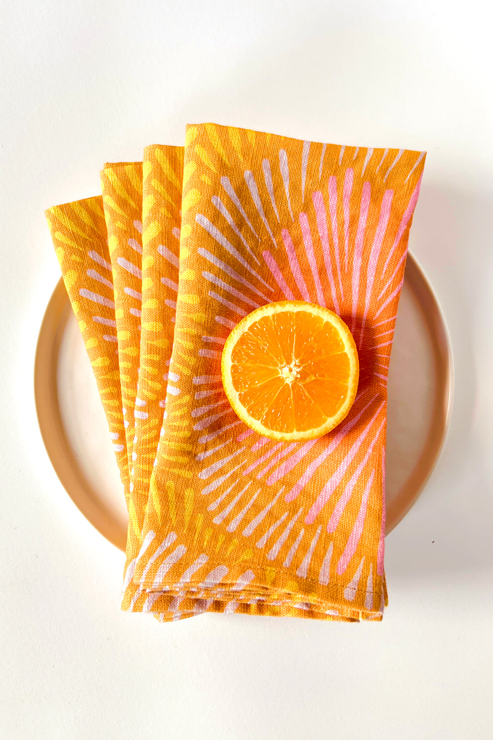 Set of four orange sun burst patterned napkins with a slice of orange on a white plate. Designed by artist Leila Simon Hayes. 