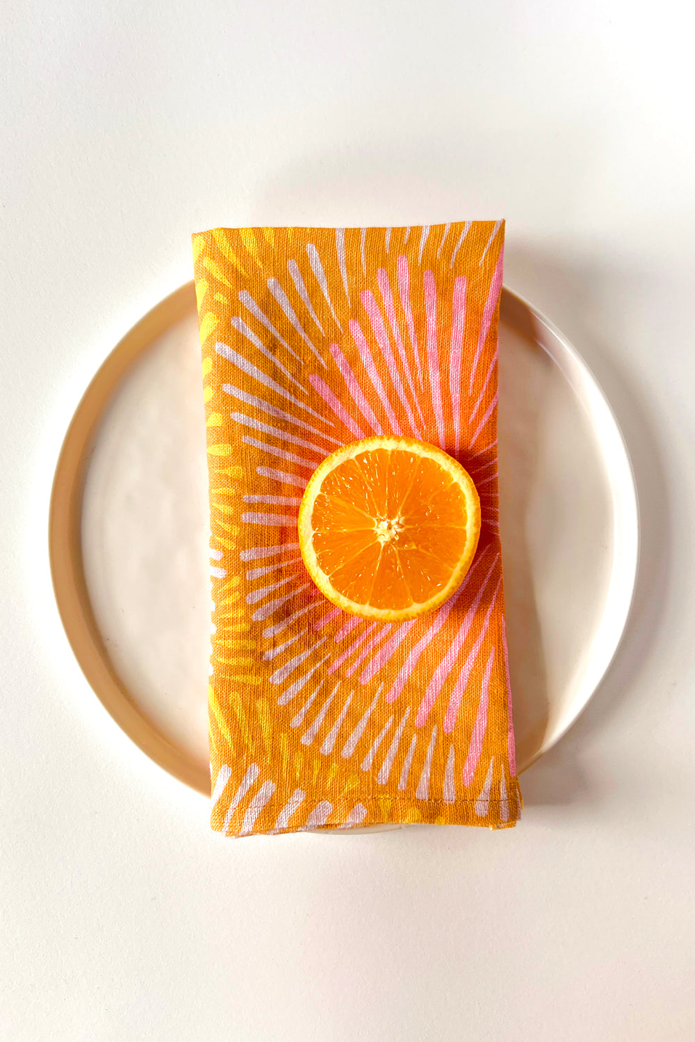 Napkin with orange sun burst design on a plate against a white background. Designed by artist Leila Simon Hayes. 