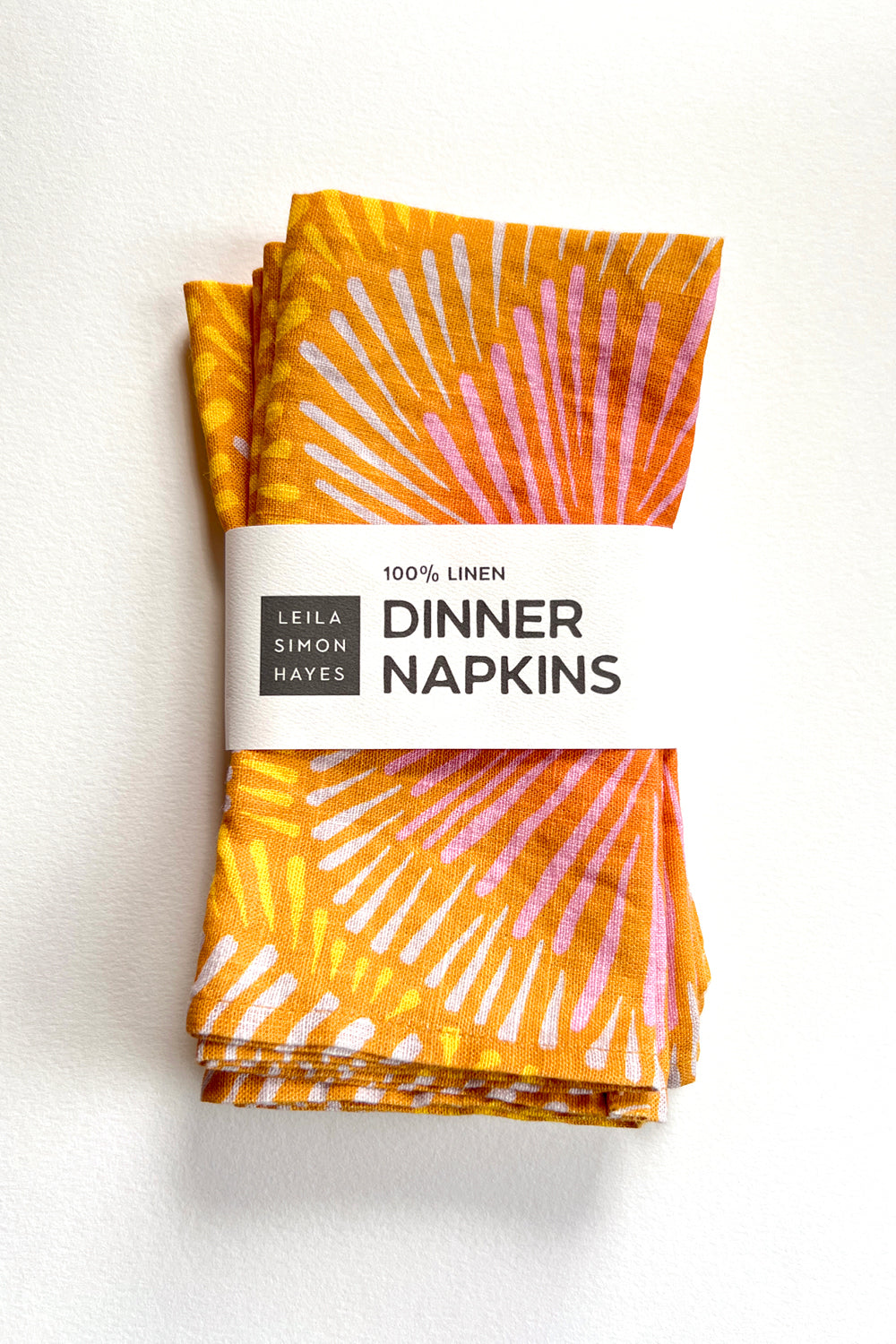 Pack of four orange dinner napkins with a sun burst pattern on a white background. The white label says "100% Linen Dinner Napkins", designed by artist Leila Simon Hayes. 