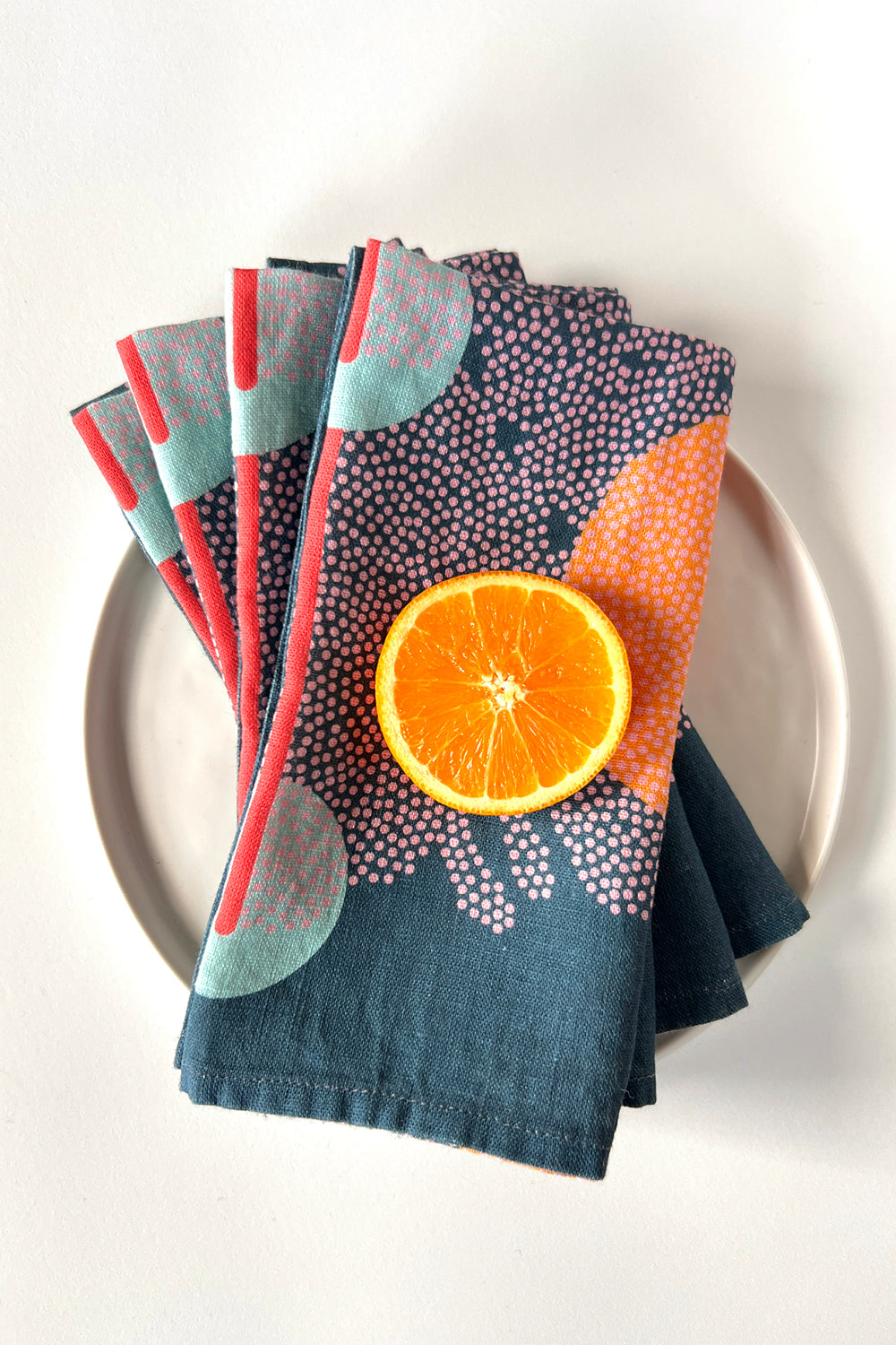 Set of four navy blue abstract patterned napkins with an orange on a white plate. Designed by artist Leila Simon Hayes. 