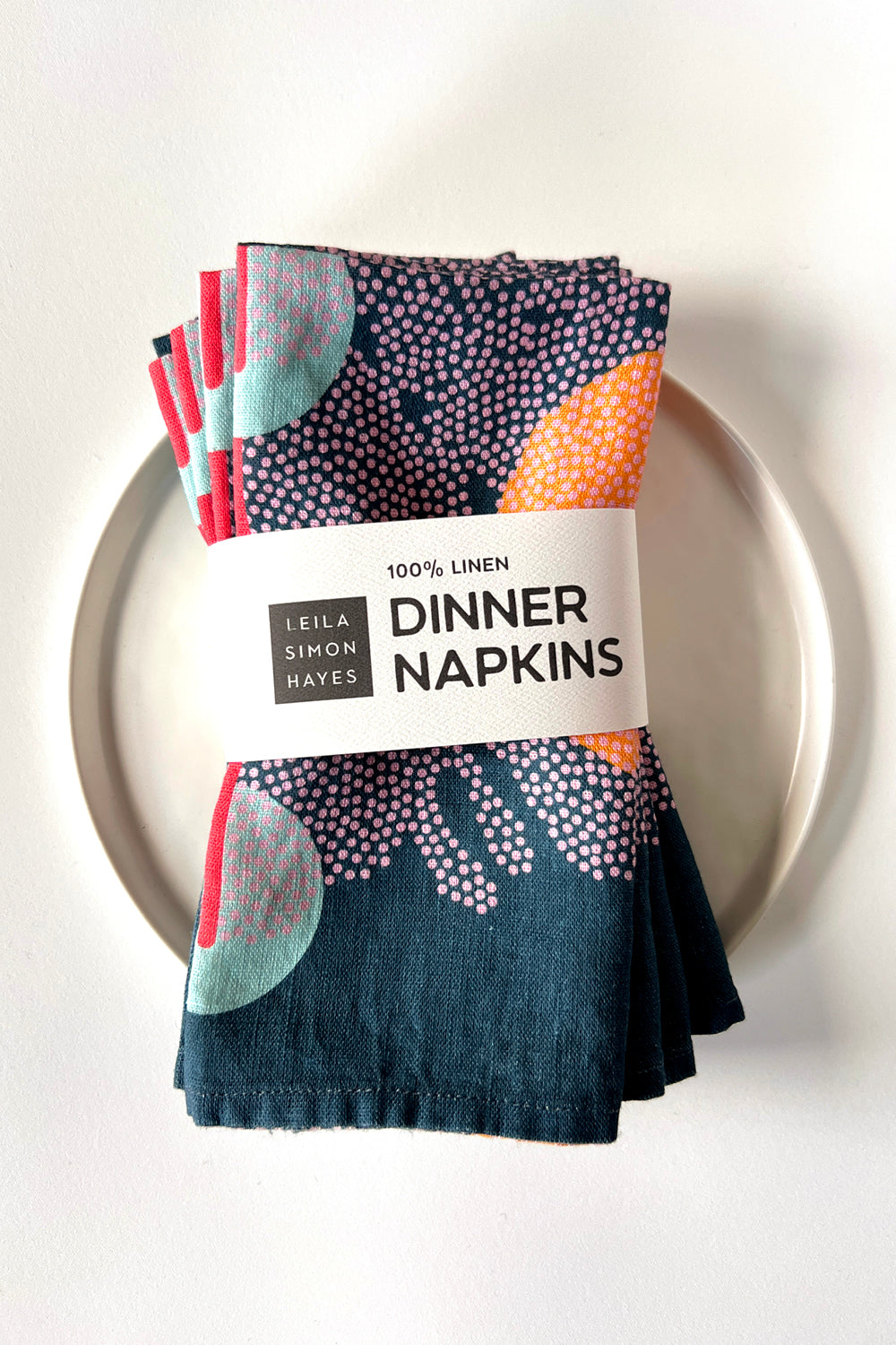 Set of four patterned navy blue dinner napkins on a white plate with a label saying "100% Linen Dinner Napkins", designed by artist Leila Simon Hayes. 