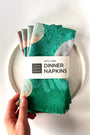 Pack of four green patterned dinner napkins with a brand label saying "100% Linen Dinner Napkins", designed by artist Leila Simon Hayes. 