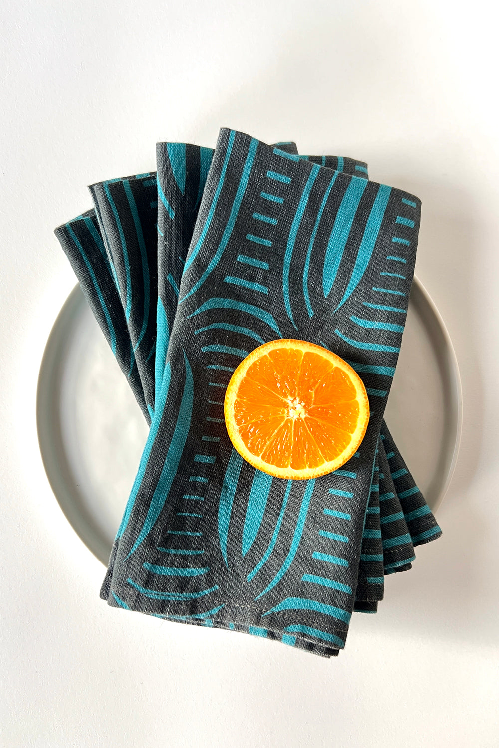 Stack of four blue stripe patterned napkins with a sliced orange on a white plate. Designed by artist Leila Simon Hayes