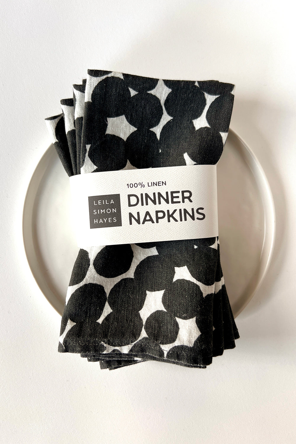 A four pack of black and white polka dot patterned dinner napkins on a white plate with a label saying "100% Linen Dinner Napkins", designed by artist Leila Simon Hayes.