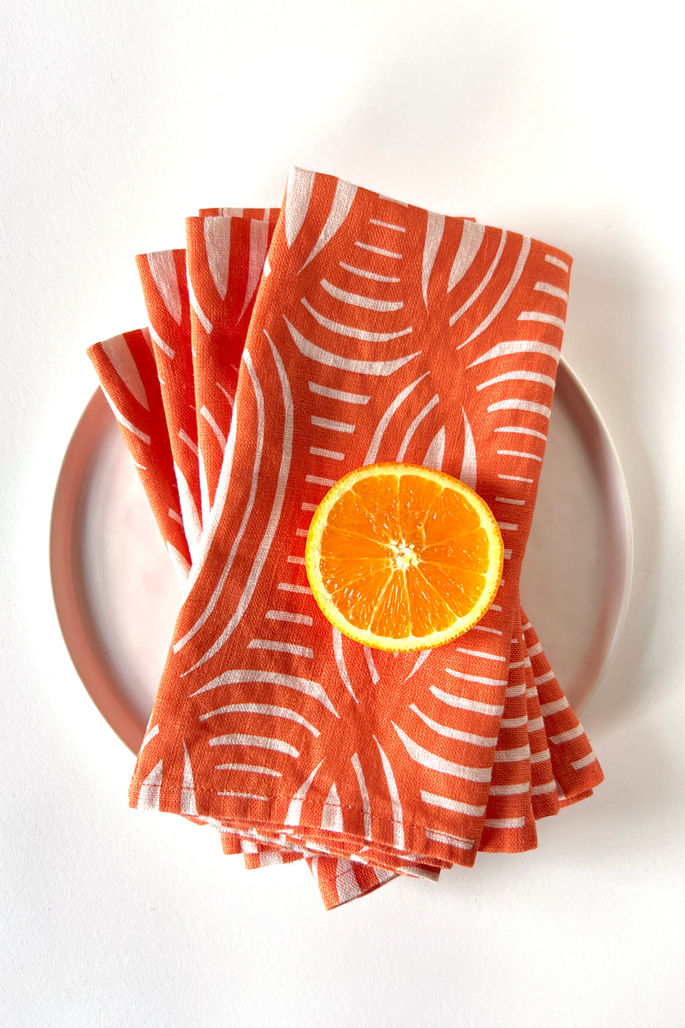 Stack of 4 coral and white patterned napkins with a sliced orange on a white plate. Designed by artist Leila Simon Hayes