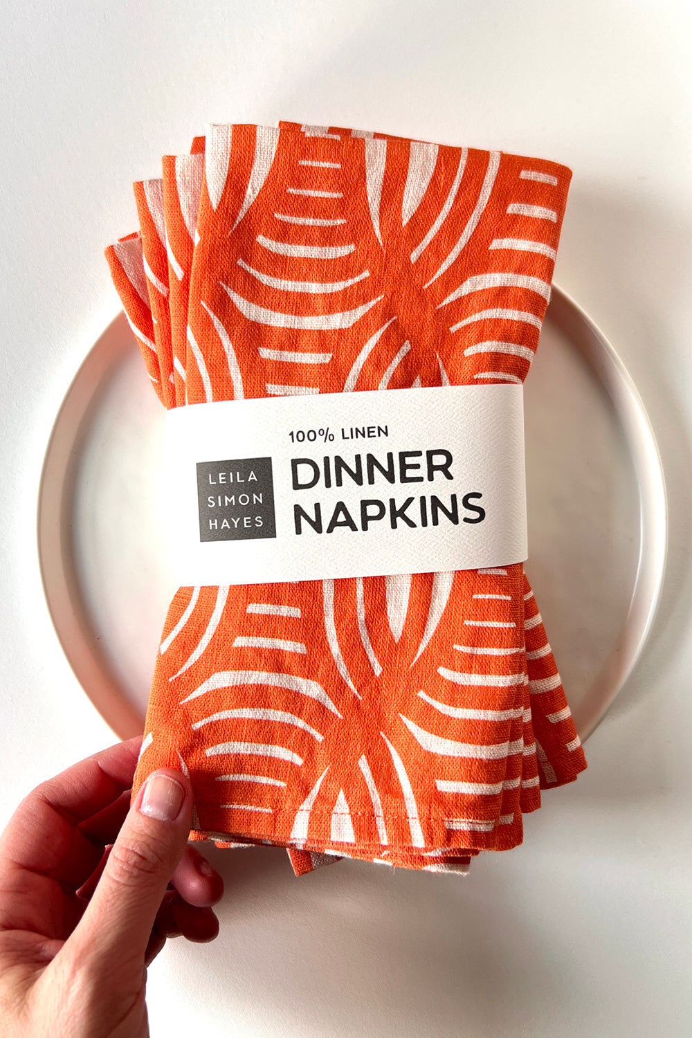 Pack of orange and white stripe patterned dinner napkins held by a hand on a white plate. Held together by a label saying "100% Linen Dinner Napkins" designed by artist Leila Simon Hayes