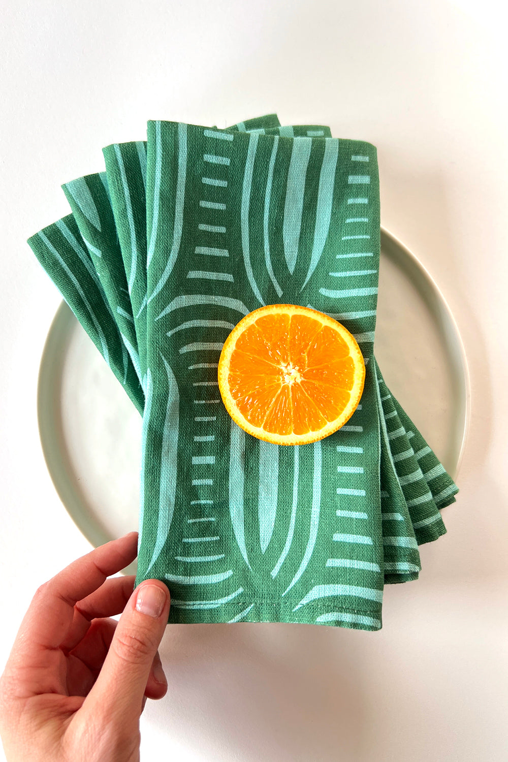 Set of 4 green striped patterned napkins layered with an orange slice on a white plate. Designed by artist Leila Simon Hayes