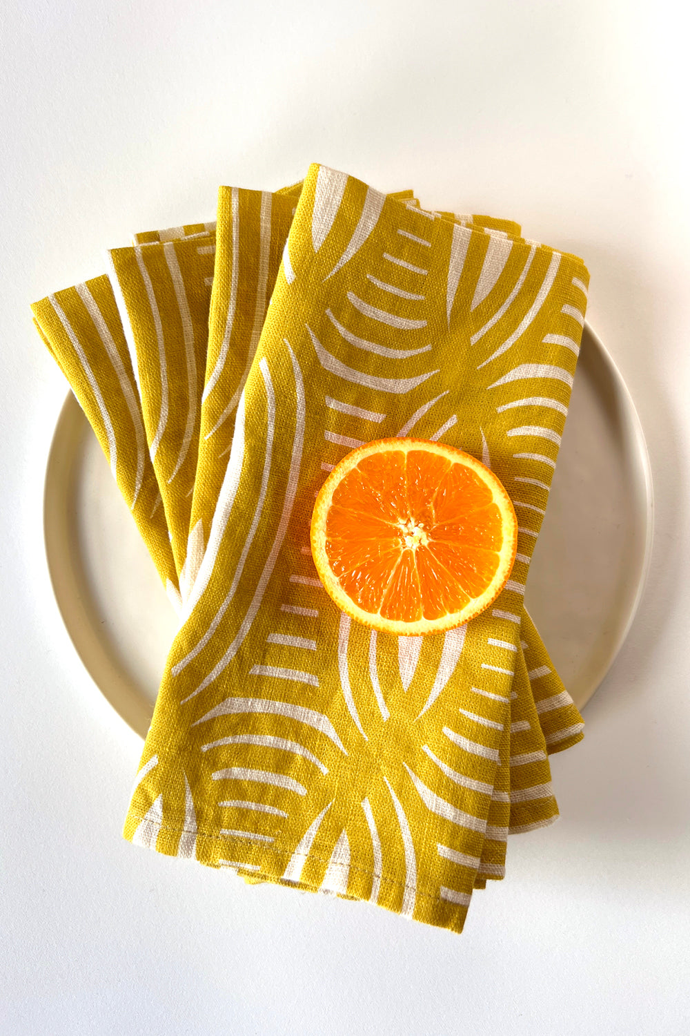 Set of four yellow patterned napkins layered over each other with a halved orange on a white plate. Designed by artist Leila Simon Hayes