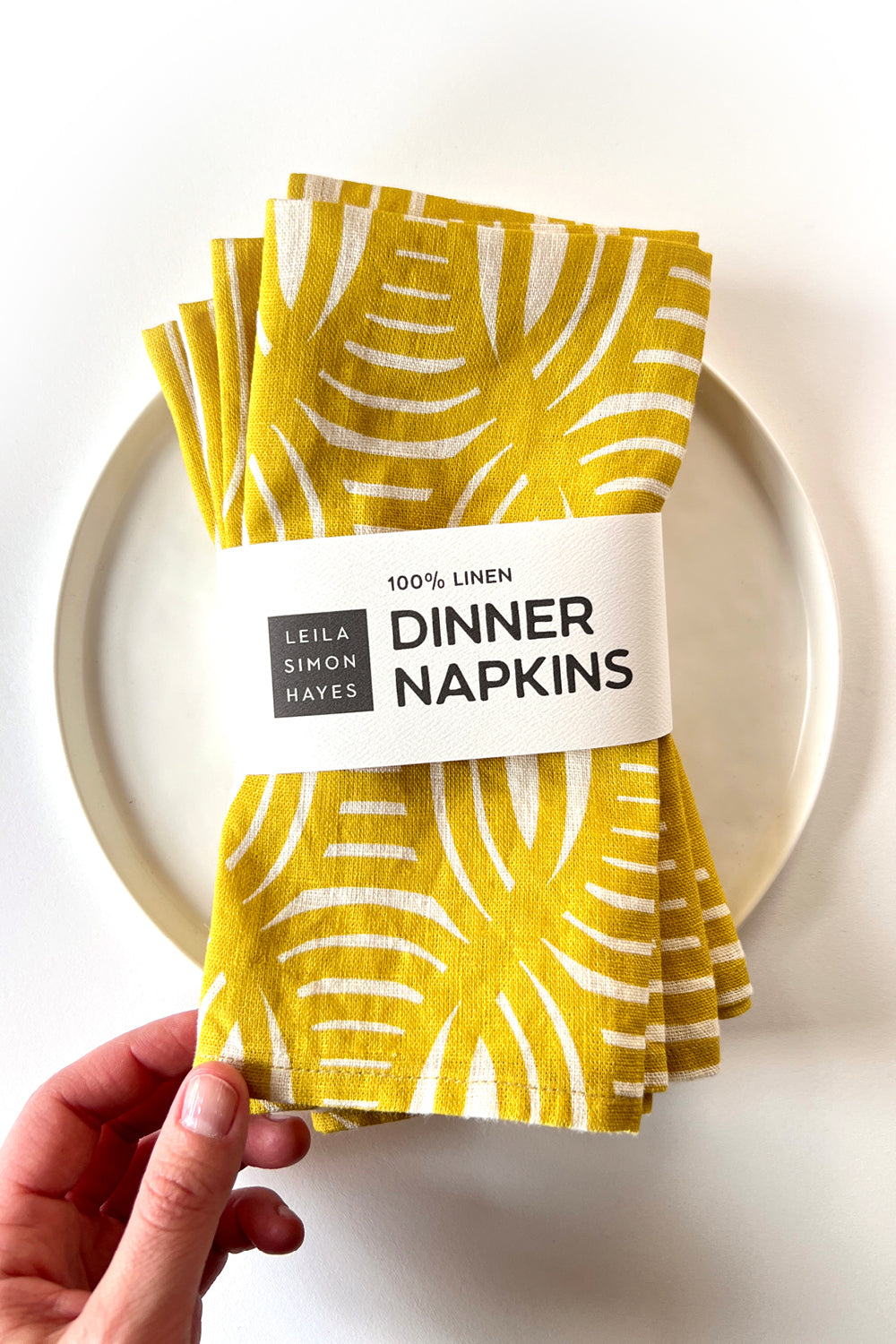 Yellow linen dinner napkins with geometrical stripe pattern held by a hand on a white plate. Designed by artist Leila Simon Hayes