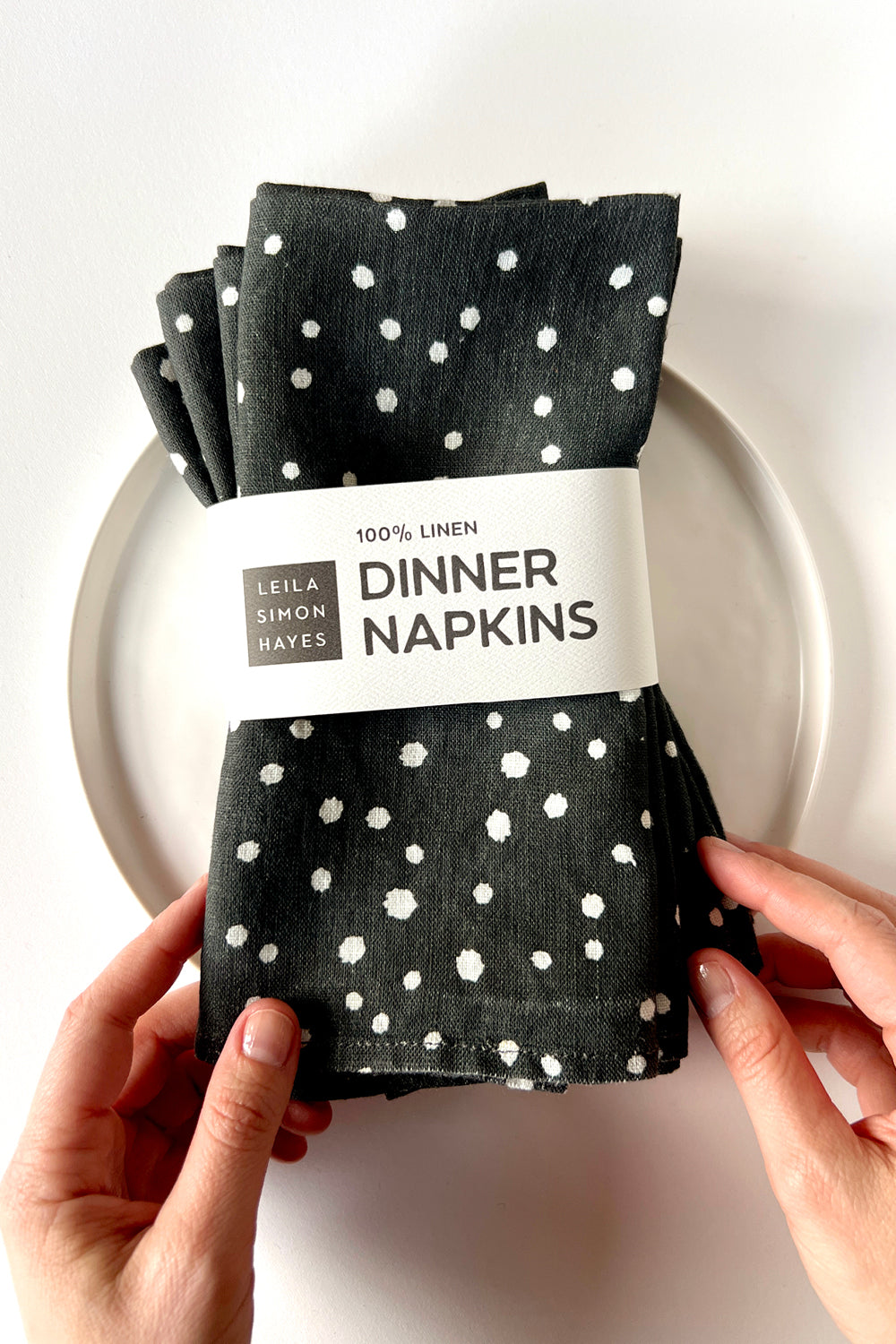 A pack of four black dinner napkins with white polka dots held by hands on a white background. The napkins are wrapped in a label saying "100% Linen Dinner Napkins", designed by artist Leila Simon Hayes
