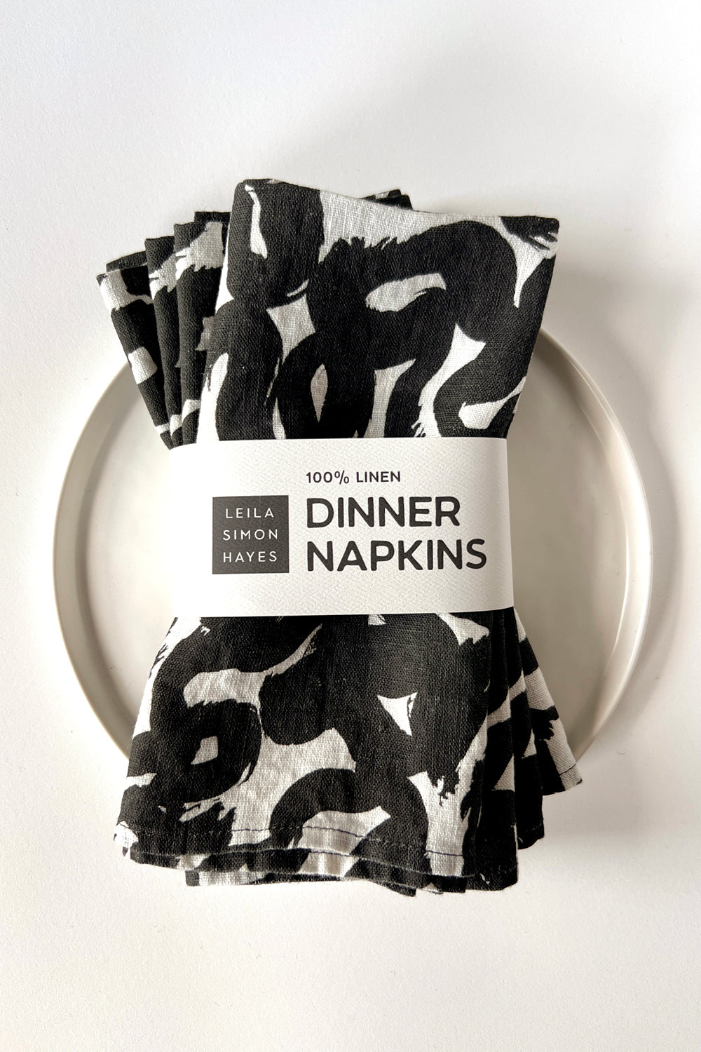 Set of four black and white patterned dinner napkins on a white plate with a label saying "100% Linen Dinner Napkins", designed by artist Leila Simon Hayes.