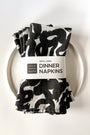 Set of four black and white patterned dinner napkins on a white plate with a label saying "100% Linen Dinner Napkins", designed by artist Leila Simon Hayes.