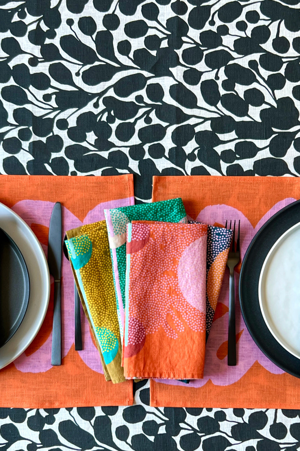 Colorful table setting with four colorful dinner napkins, pink and coral patterned placemats and a black and white floral tablecloth. Designed by artist Leila Simon Hayes.