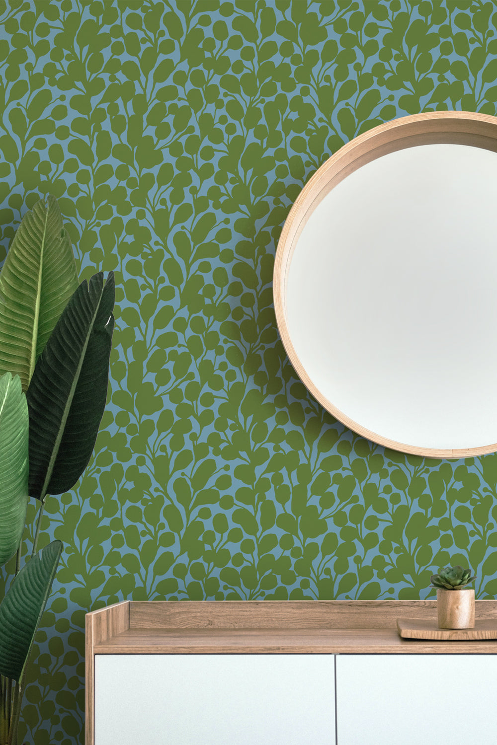 Green and blue botanical patterned wallpaper shown in a styled interior setting with a round wooden-framed mirror, a light wood console table, white cabinet doors, and a potted plant. Designed by artist Leila Simon Hayes.
