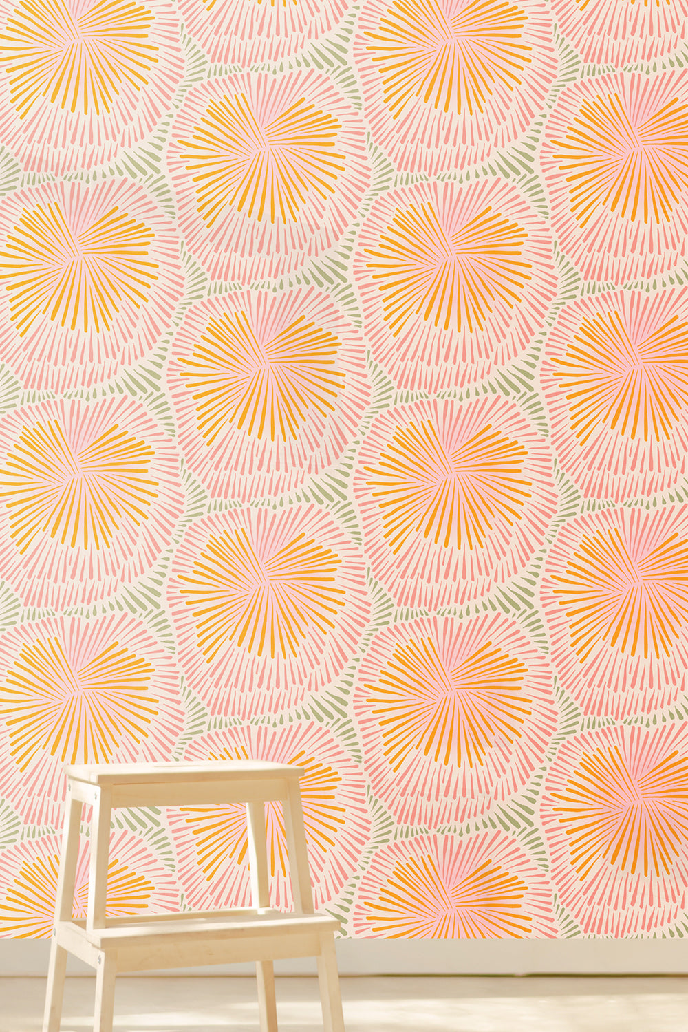 A wall covered in a repeating botanical pattern wallpaper with pink, orange, and light green palm-like motifs on a white background, with a small light wood stool in the foreground. Designed by artist Leila Simon Hayes.
