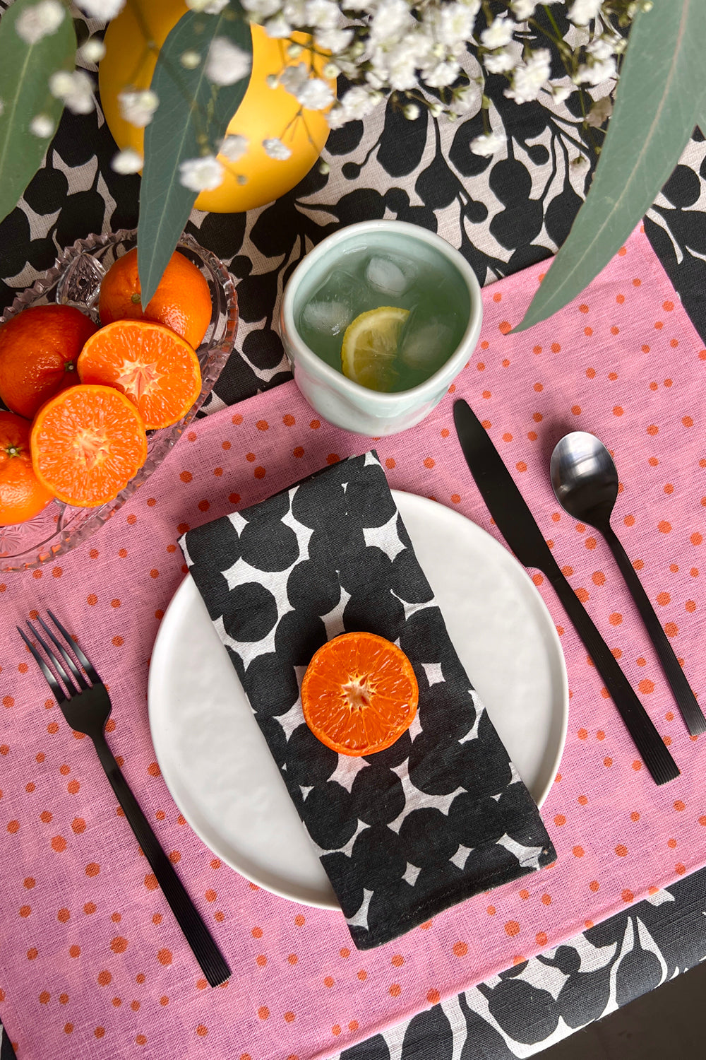 Table setting with a black and white dots patterned napkin, pink placemat with orange organic dots, and black and white botanical tablecloth. Designed by artist Leila Simon Hayes