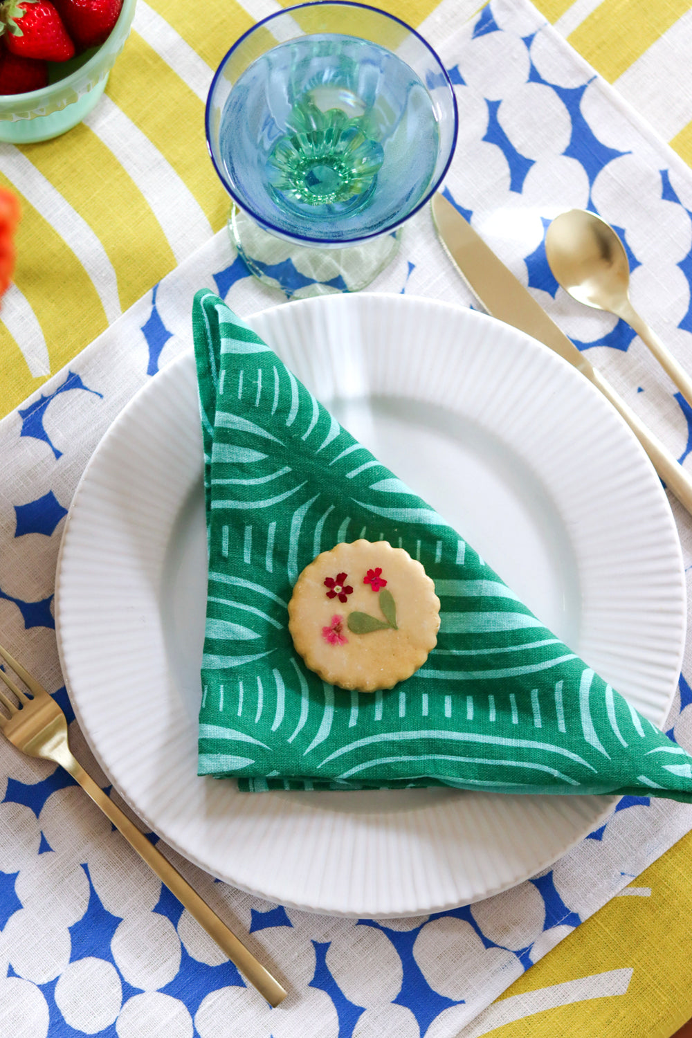 Table setting with a green stripe patterned napkin, blue and white dotted placemat, and yellow striped tablecloth. A green linen napkin with a striped geometric pattern, a slice of orange placed on top of it. Designed by artist Leila Simon Hayes
