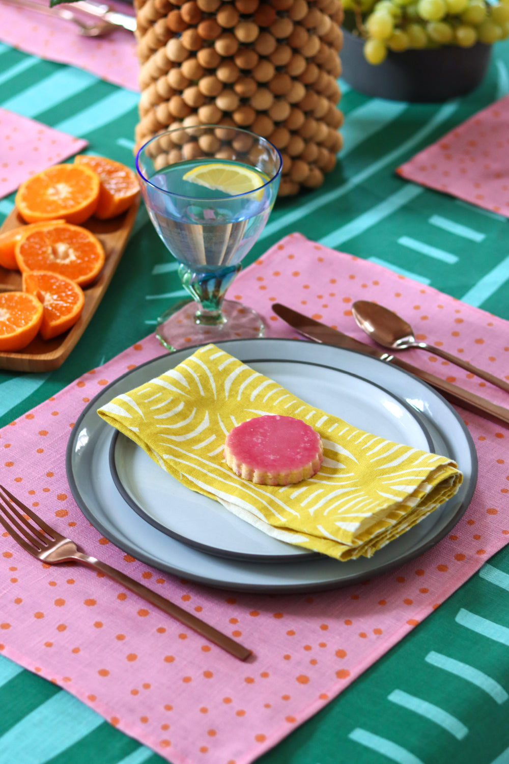 Table setting with a yellow striped napkin design, a pink linen placemat, and a green striped tablecloth. Designed by artist Leila Simon Hayes
