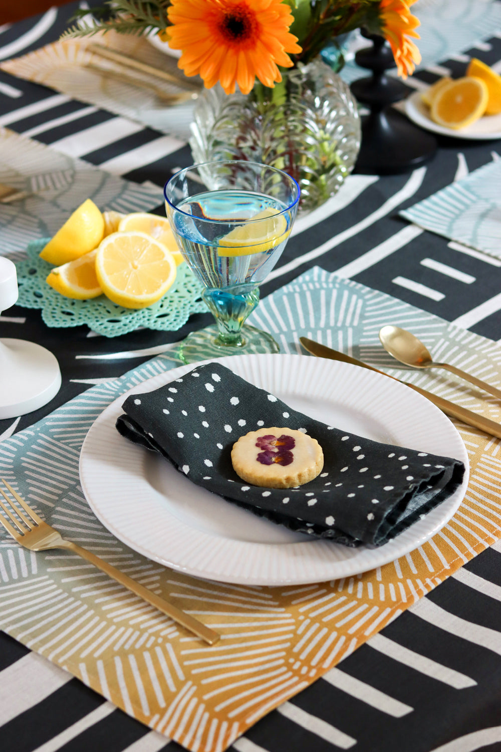 Dining table setting with black and white small dot napkins, a blue and yellow placemat, over a black and white geometric tablecloth. Designed by artist Leila Simon Hayes.