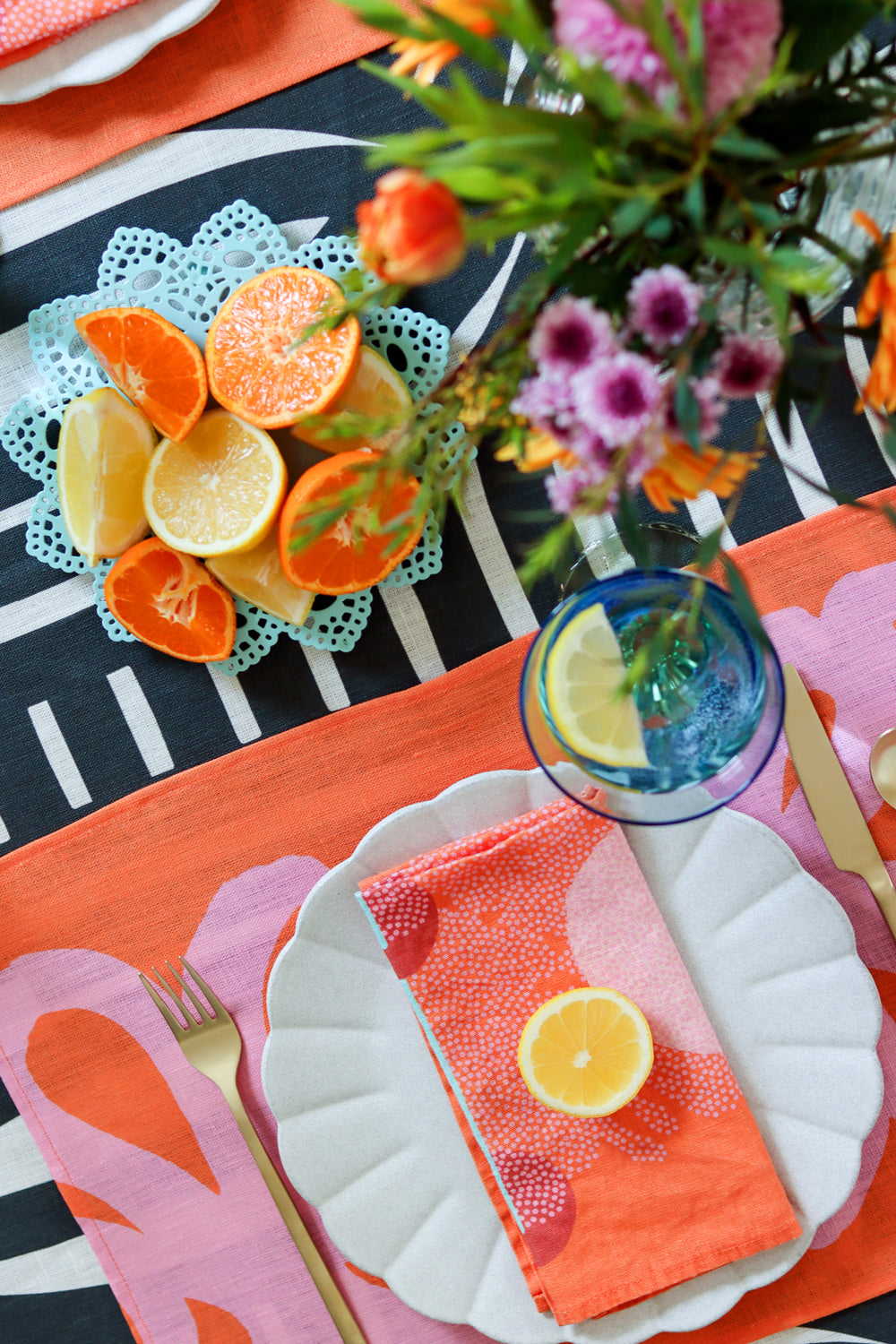 Dining table setting with red napkins, coral and pink placemats, and a black and white striped tablecloth with a vase of flowers. Table linens designed by artist Leila Simon Hayes