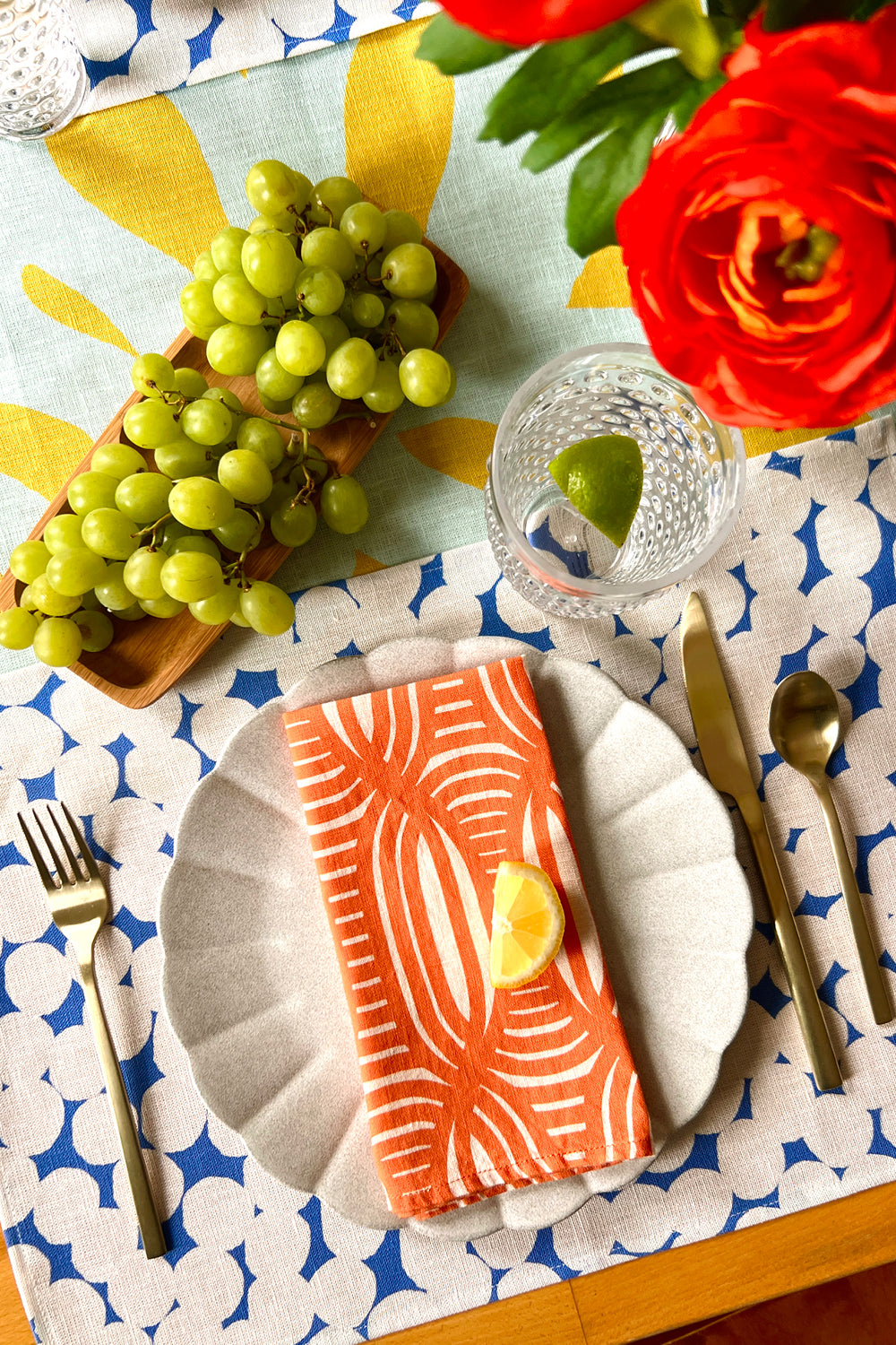 Dining table setting with a coral linen napkin, blue and white dots placemat, over a citron abstract table runner. Designed by artist Leila Simon Hayes
