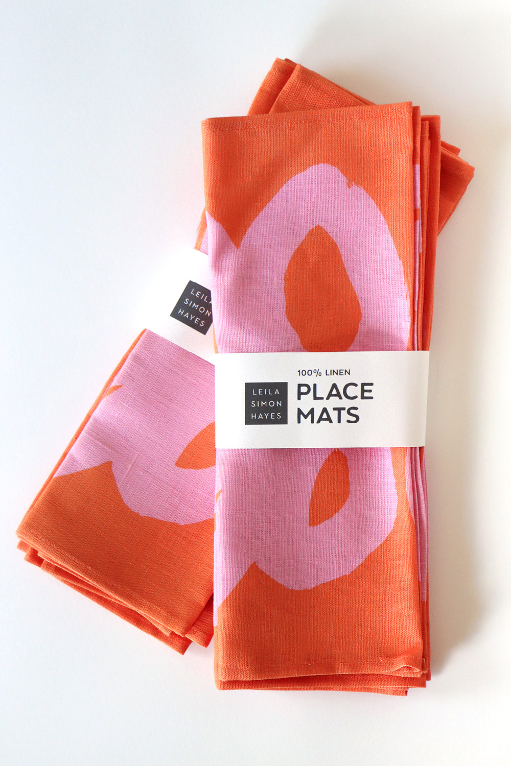 Orange place mats with pink floral design on a white background. A label wrapped around the placemats says "100% linen placemats" designed by artist Leila Simon Hayes
