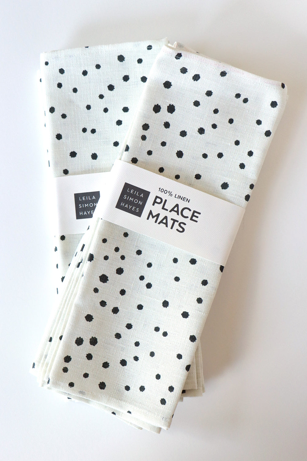 A white linen placemat with a black organic dotted pattern, with a label that says "100% Linen Placemats" designed by artist Leila Simon Hayes