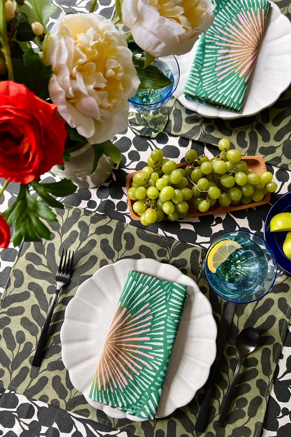 Decorative table setting with green burst napkins, olive green floral placemats, and a black and white botanical tablecloth.